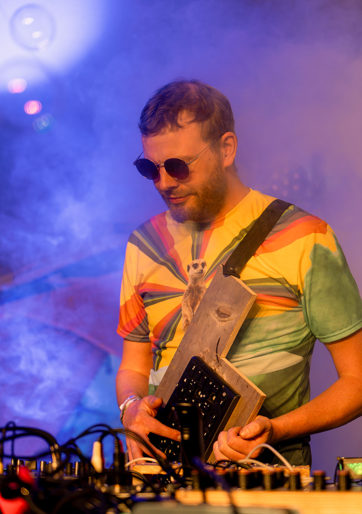 Foerb mit Sonnenbrille an seinen selbstgebauten Geräten, eine Tastatur auf einem Holzbrett umgehängt, im blauen Bühnennebel